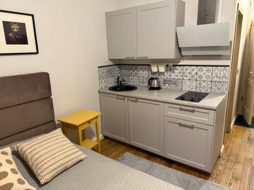 A small kitchen area with cabinets, a sink, and a stove beside a bed with decorative pillows.