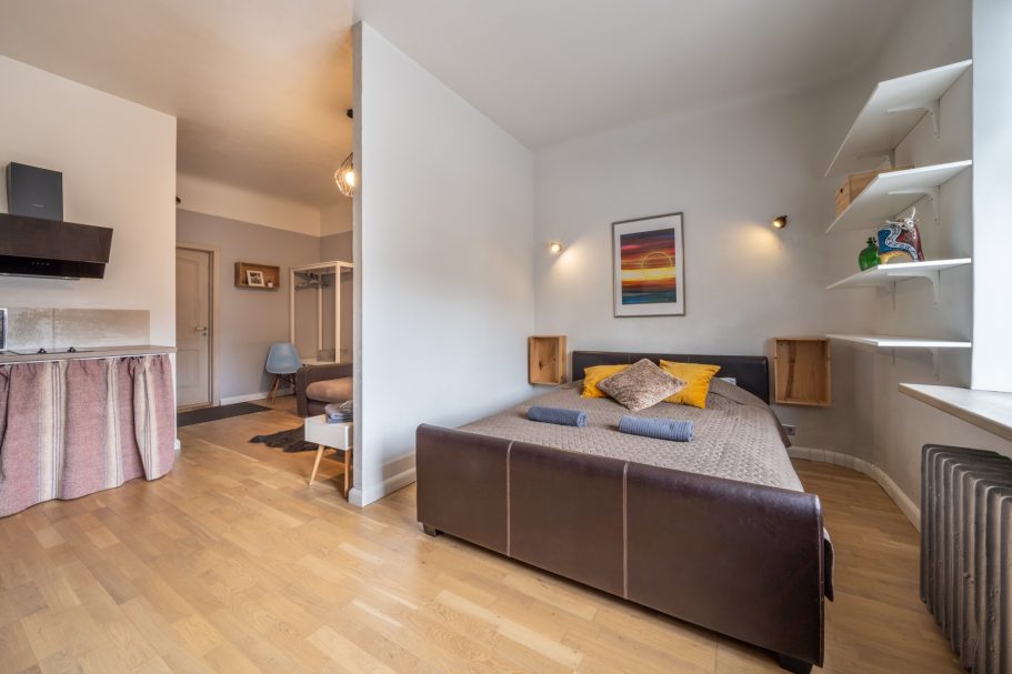 Cozy studio apartment with a bed, wooden floors, and shelves, featuring warm decor.