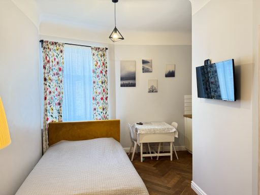 Cozy bedroom with a bed, TV, and a small desk near a window with patterned curtains.