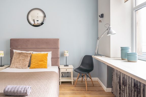 Cozy bedroom with a bed, chair, desk, and light blue walls. Decor includes a round wall clock.