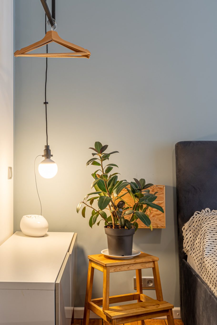 Cozy bedroom corner with hanging light, plant, and wooden stool beside a nightstand.