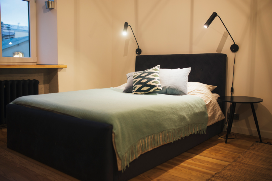 Modern bedroom with a dark bed, green blanket, decorative pillows, and reading lamps.