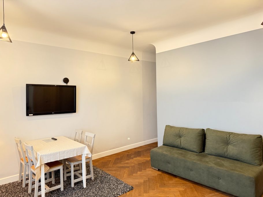 Cozy living area with a green sofa, small dining table, and a wall-mounted TV.