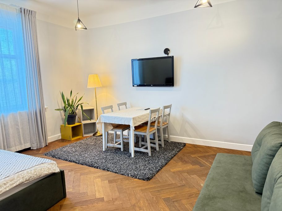 Cozy living space with a bed, dining table, and potted plant against a light wall.