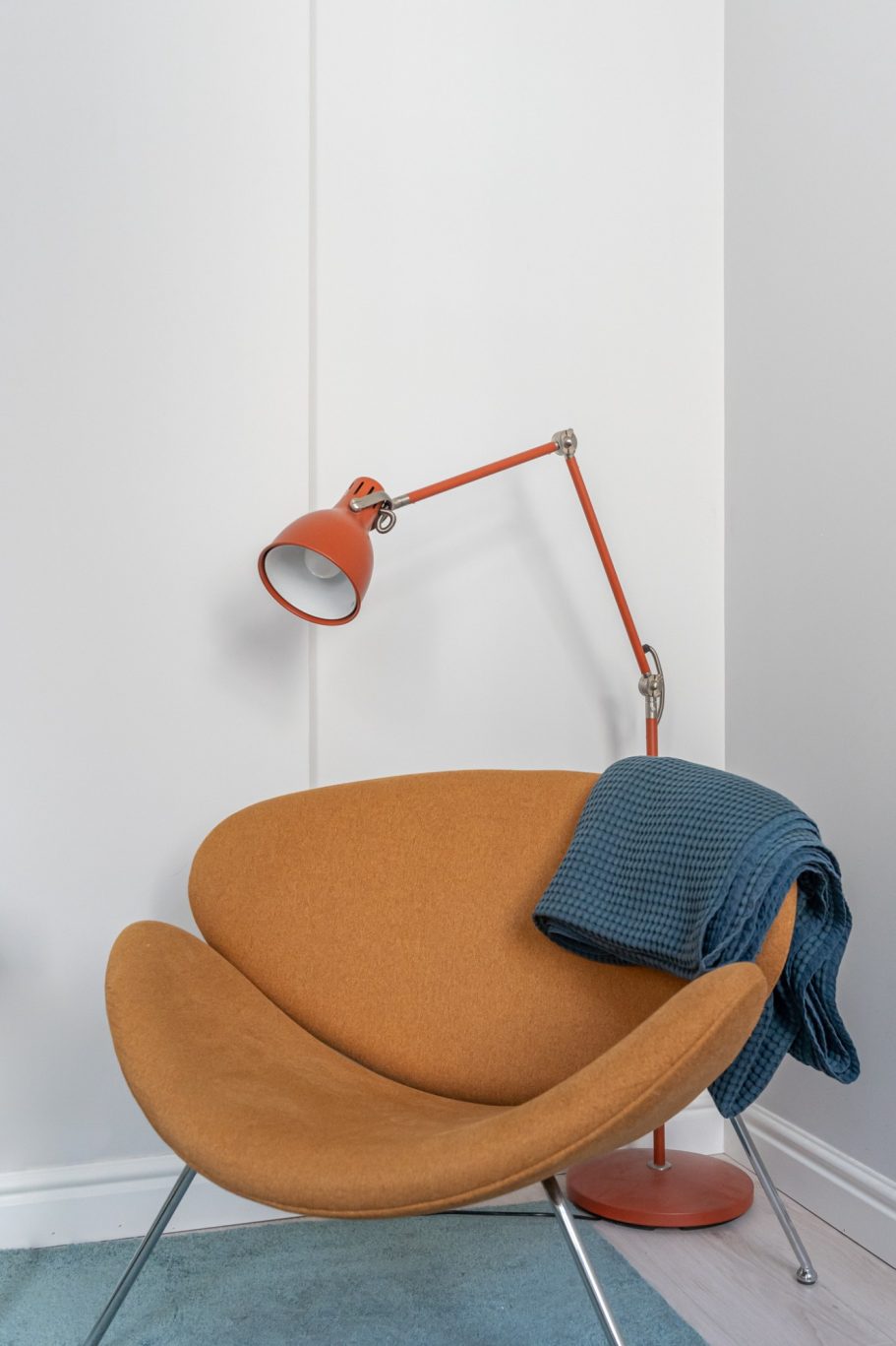 A cozy orange chair with a blue blanket and an angled red lamp in a bright room.