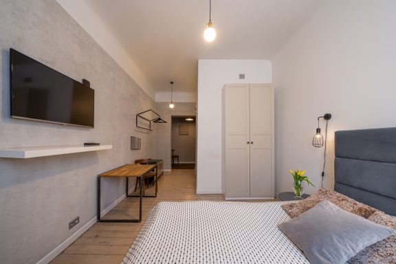 Modern bedroom with a TV, desk, and light-colored walls. A flower in a vase adds a touch of color.
